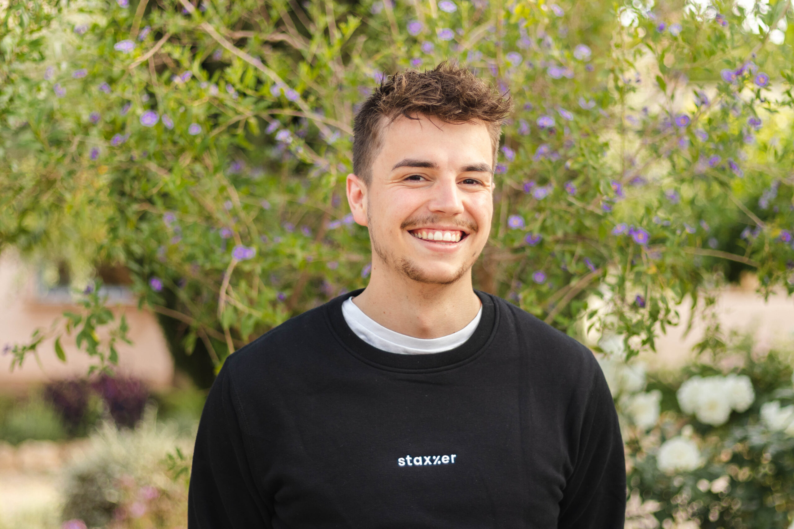 A young man in a black sweatshirt smiling in front of bushes.