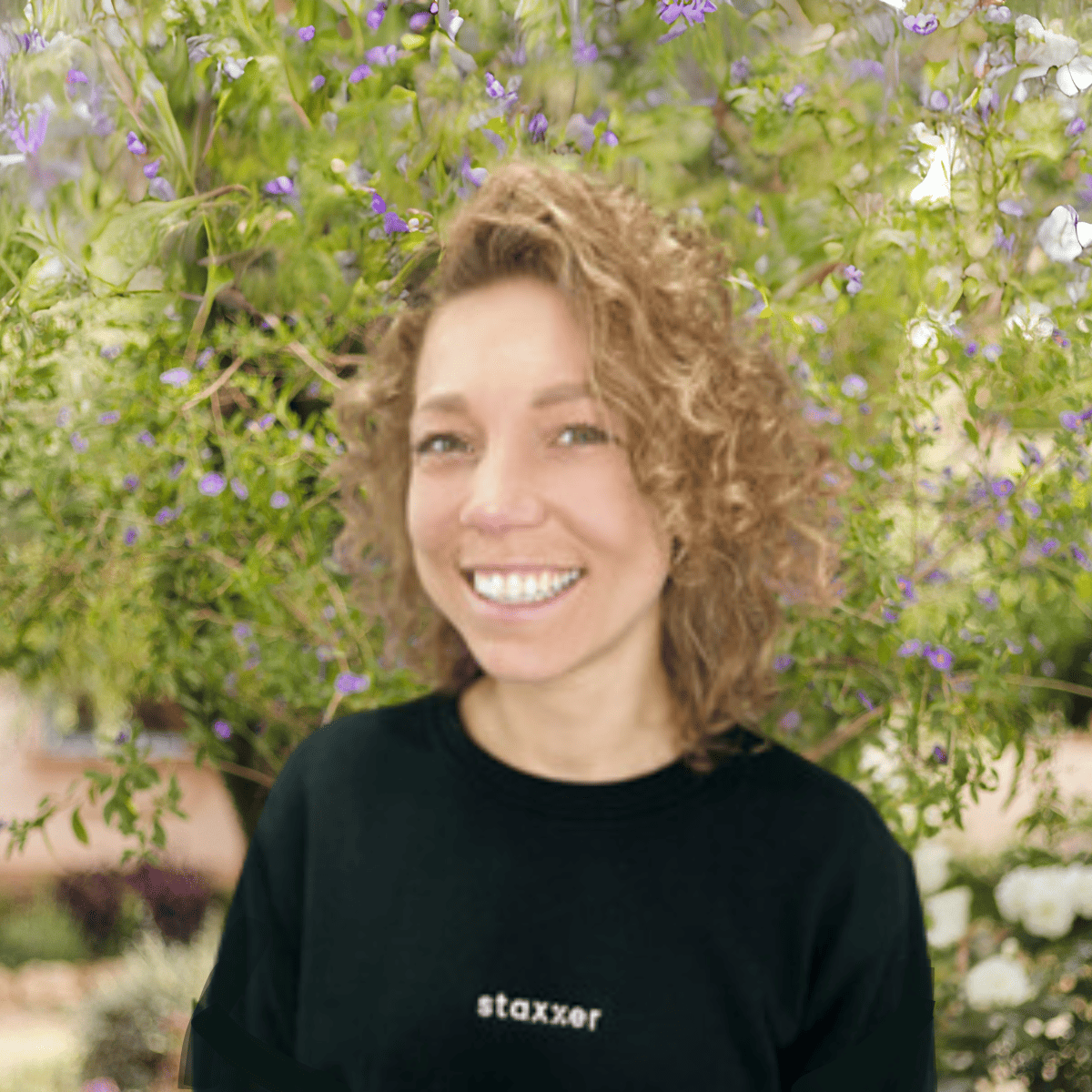 A woman with curly hair smiling among flowers.