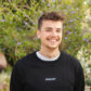 Een jonge man in een zwart sweatshirt glimlachend voor struiken.